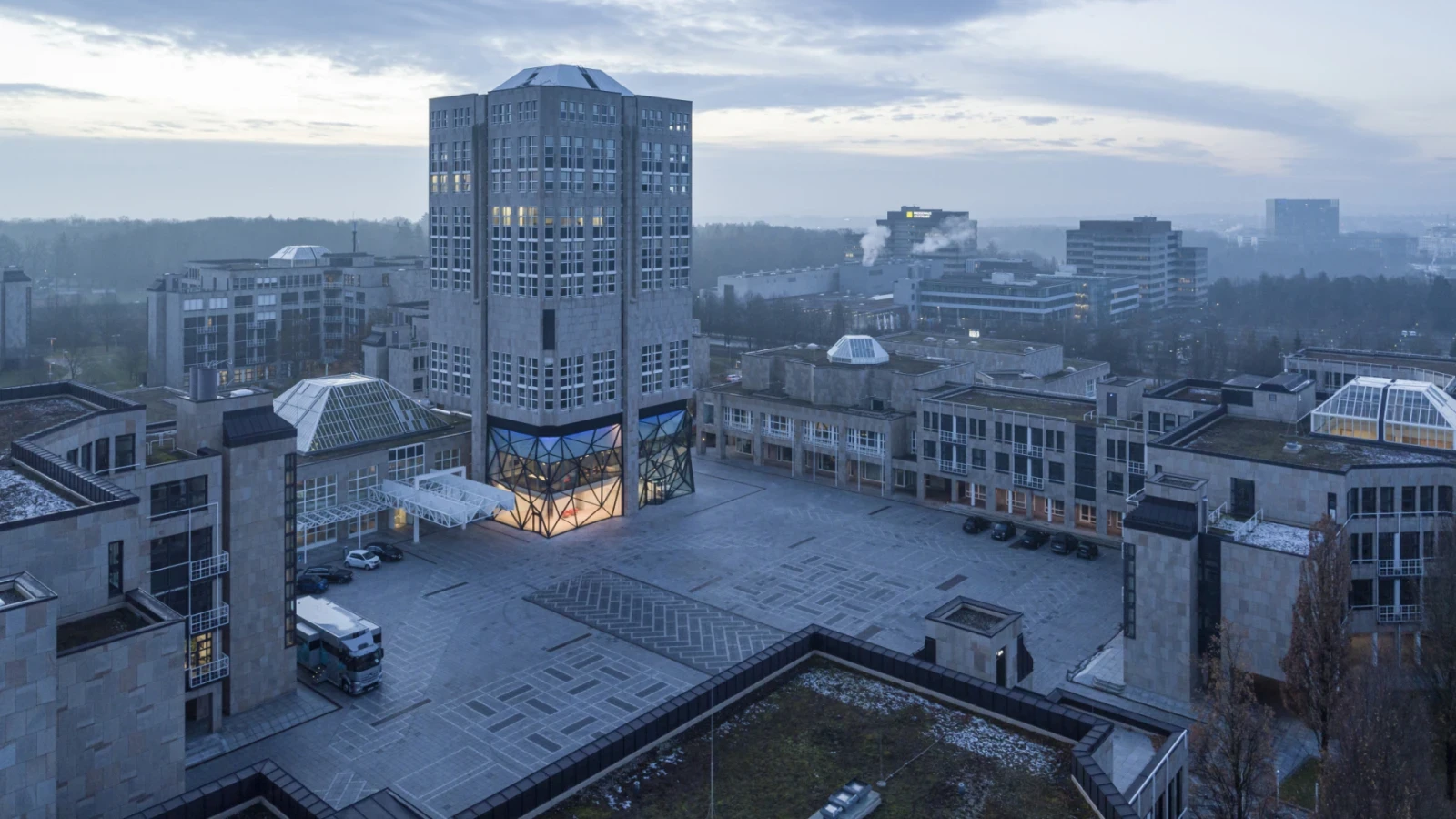 Luftaufnahme der Sternhöhe in Stuttgart in der Abenddämmerung, mit Turm, beleuchtetem Glasfoyer und großem Platz